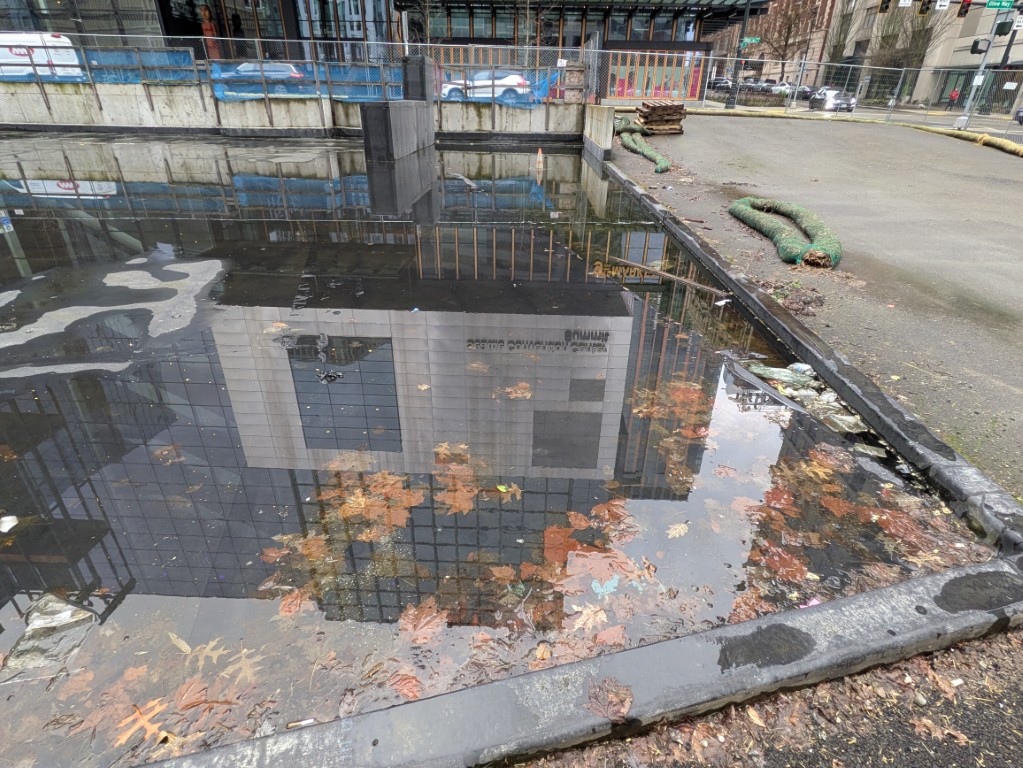 The Summit reflected upside-down in a puddle on the 920 Olive Way parcel — a fully entitled tower site, fenced and flooding.