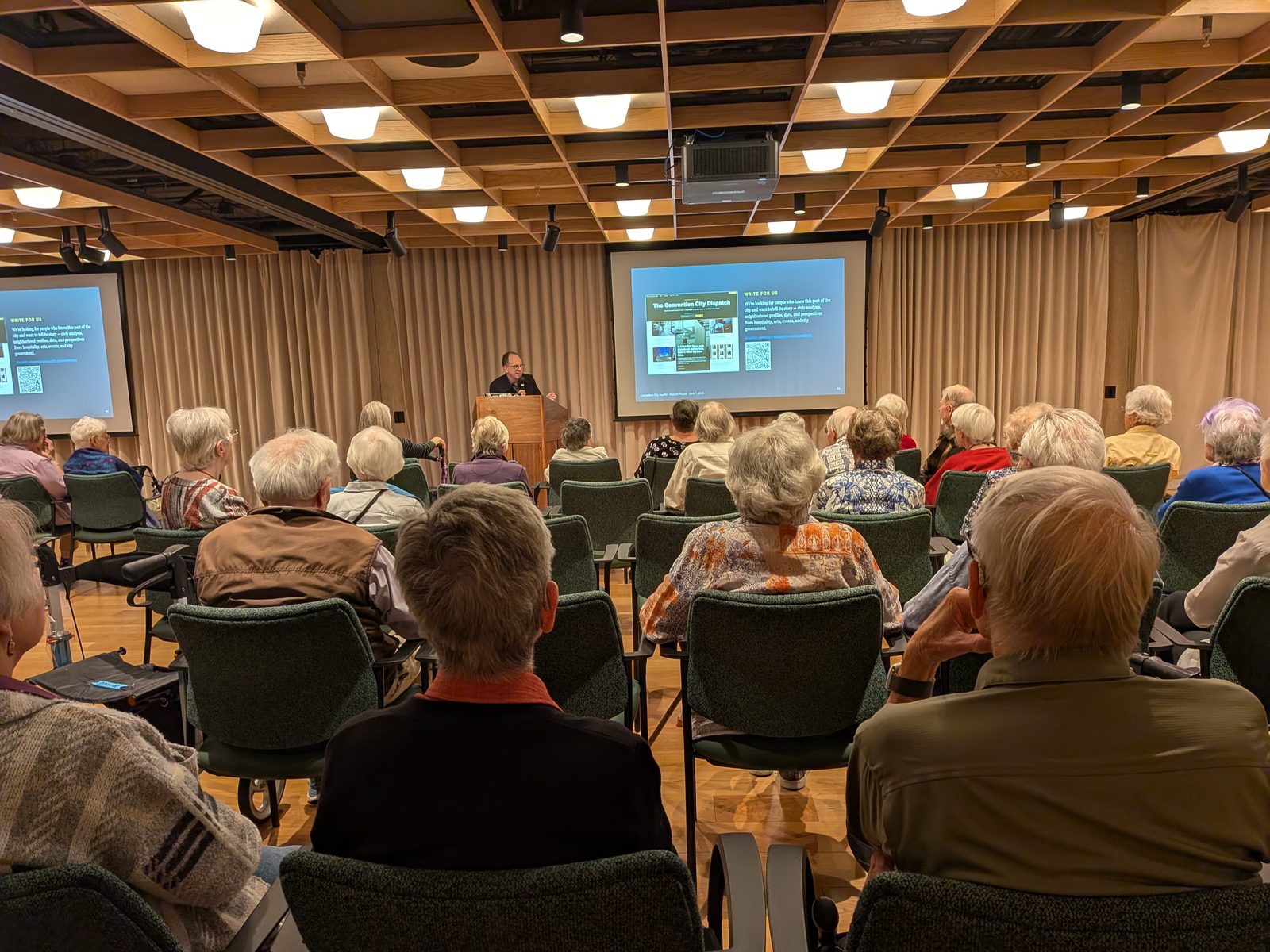 A speaker at a podium addresses a full room of seated Horizon House residents, with a "Write For Us" Convention City Dispatch slide projected on the screen.
