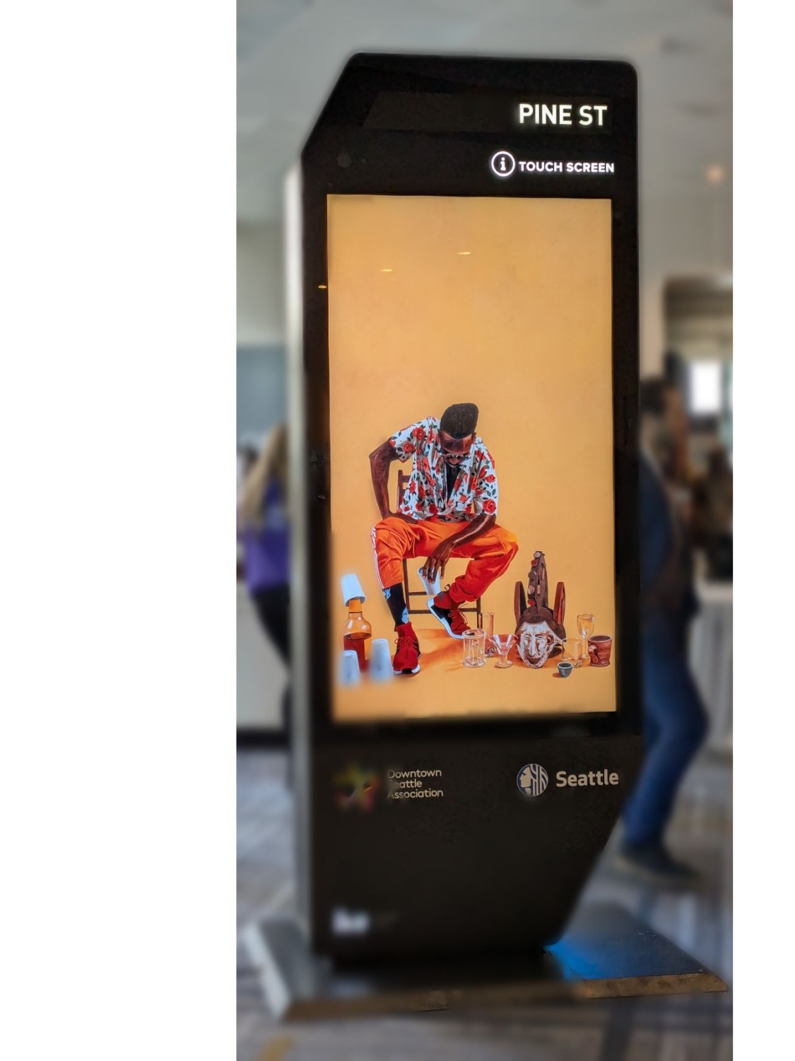 An IKE digital advertising kiosk labeled PINE ST with a touchscreen displaying an ad, Downtown Seattle Association and Seattle logos at its base, in a conference hall