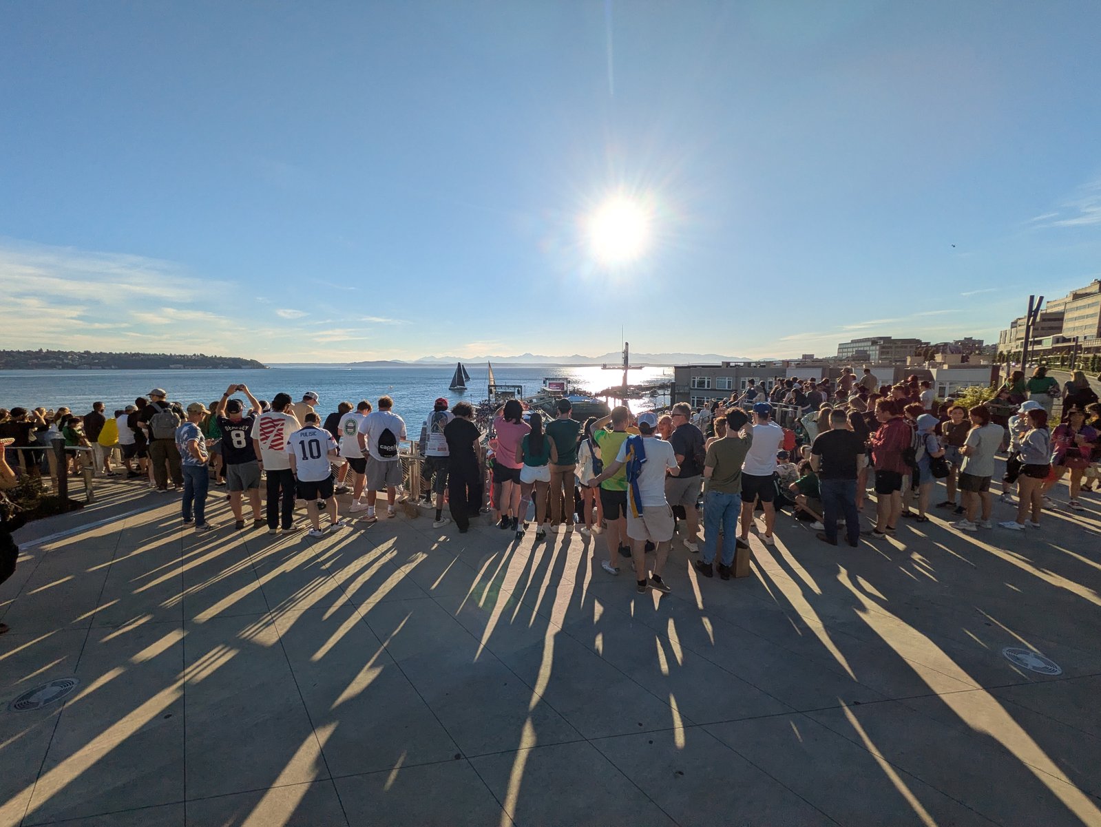 A large crowd of soccer fans in jerseys on a sunlit waterfront plaza overlooking Elliott Bay and the Olympic Mountains, long shadows across the pavement.