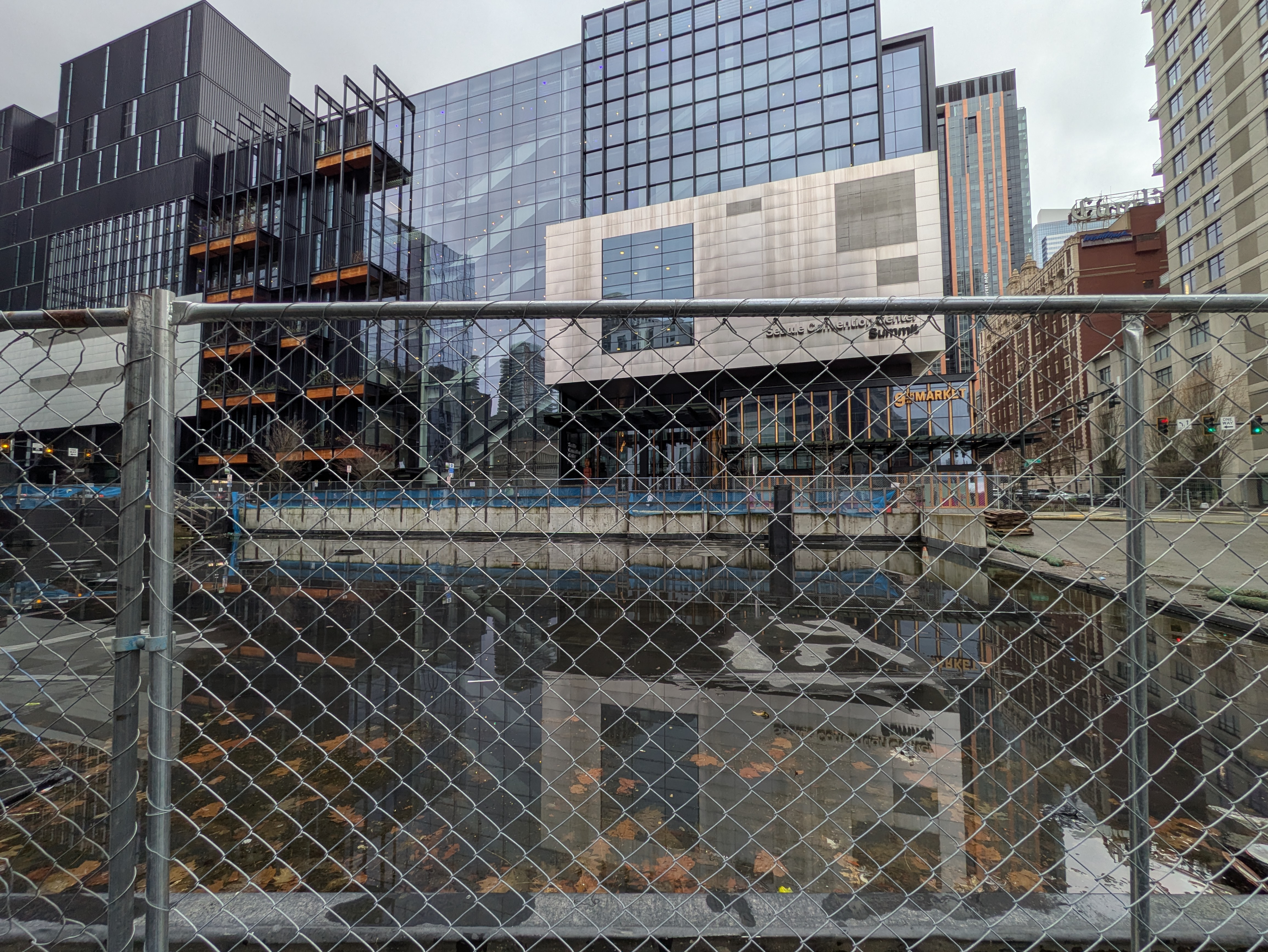 The Seattle Convention Center's Summit building seen through a chain-link construction fence, its facade reflected in a pool of standing water on the fenced-off ground in front of it.