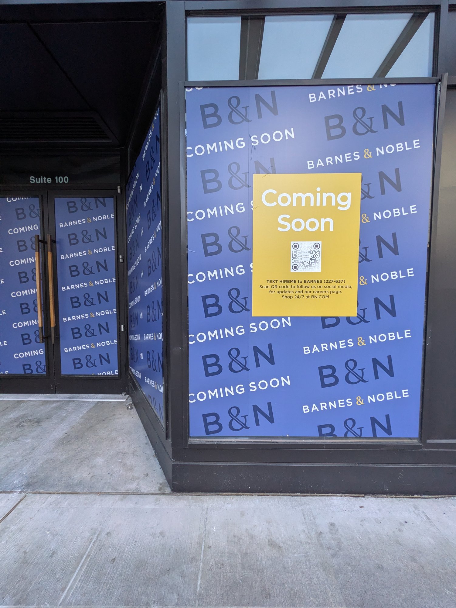 A Pike Street storefront papered floor to ceiling in blue Barnes & Noble 'Coming Soon' wraps, with a bright yellow 'Coming Soon' poster and a QR code inviting passersby to apply for jobs.