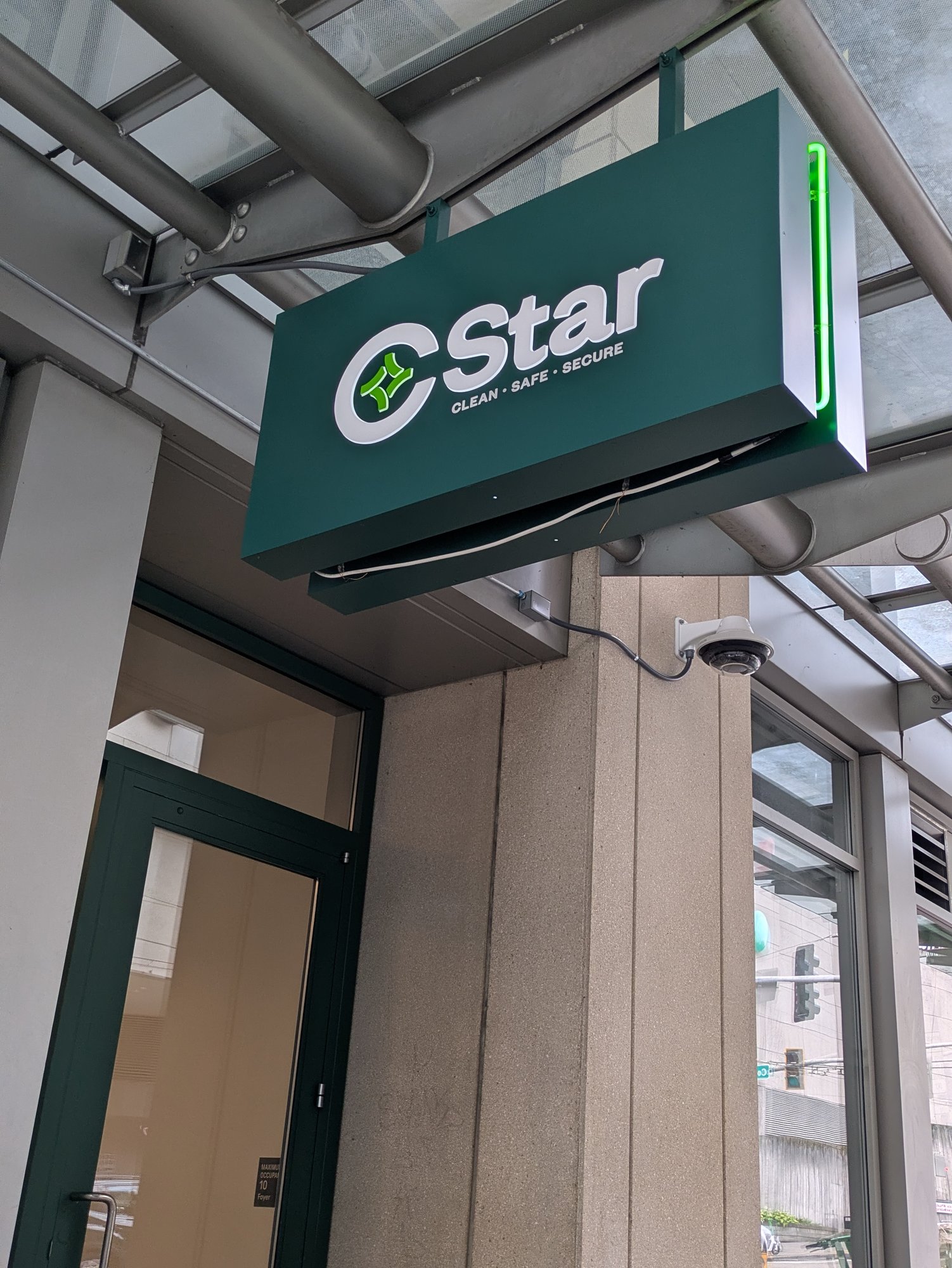 A green blade sign reading "CLEAN · SAFE · SECURE" with a "CStar" logo juts out over an empty Pike Street sidewalk, a security camera mounted just below it.