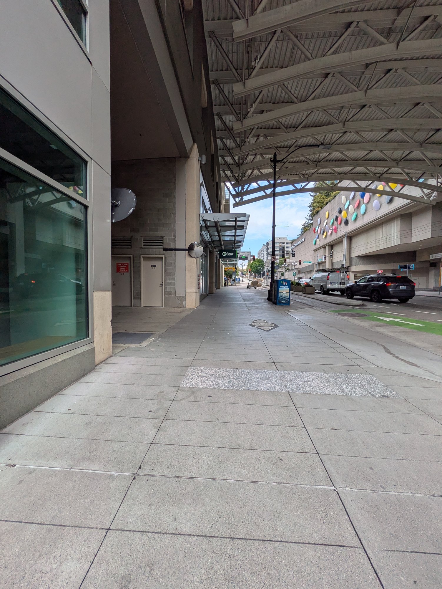 An empty sidewalk runs the length of Pike Street under the convention center's barrel-vaulted glass canopy; the small green CStar blade sign hangs in the middle distance, the convention center's dot mural on the wall across an empty street.