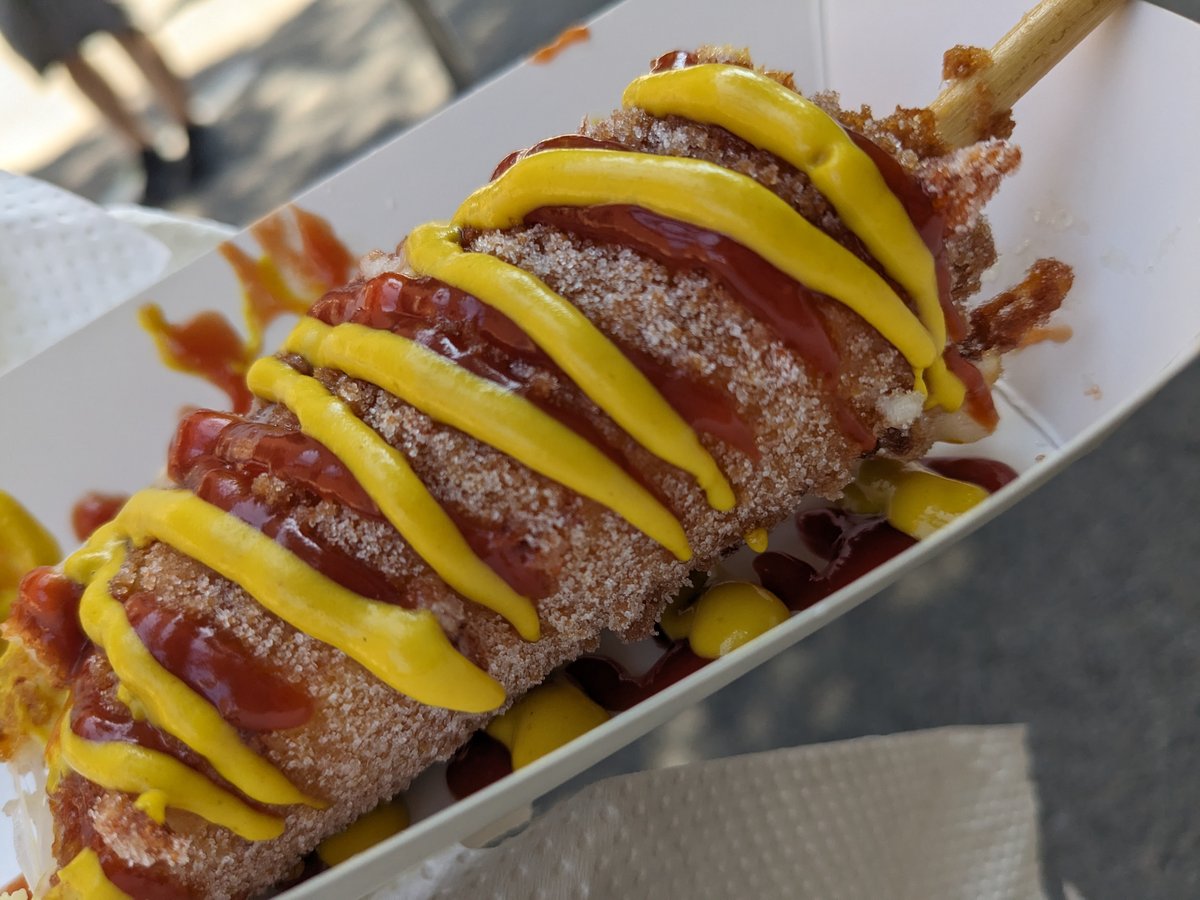 A sugar-dusted Korean corn dog on a stick, drizzled with ketchup and mustard, in a paper tray.