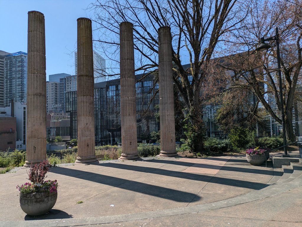 Four weathered classical columns stand in a plaza, the convention center's glass wall and downtown towers behind them, long shadows across the pavement.