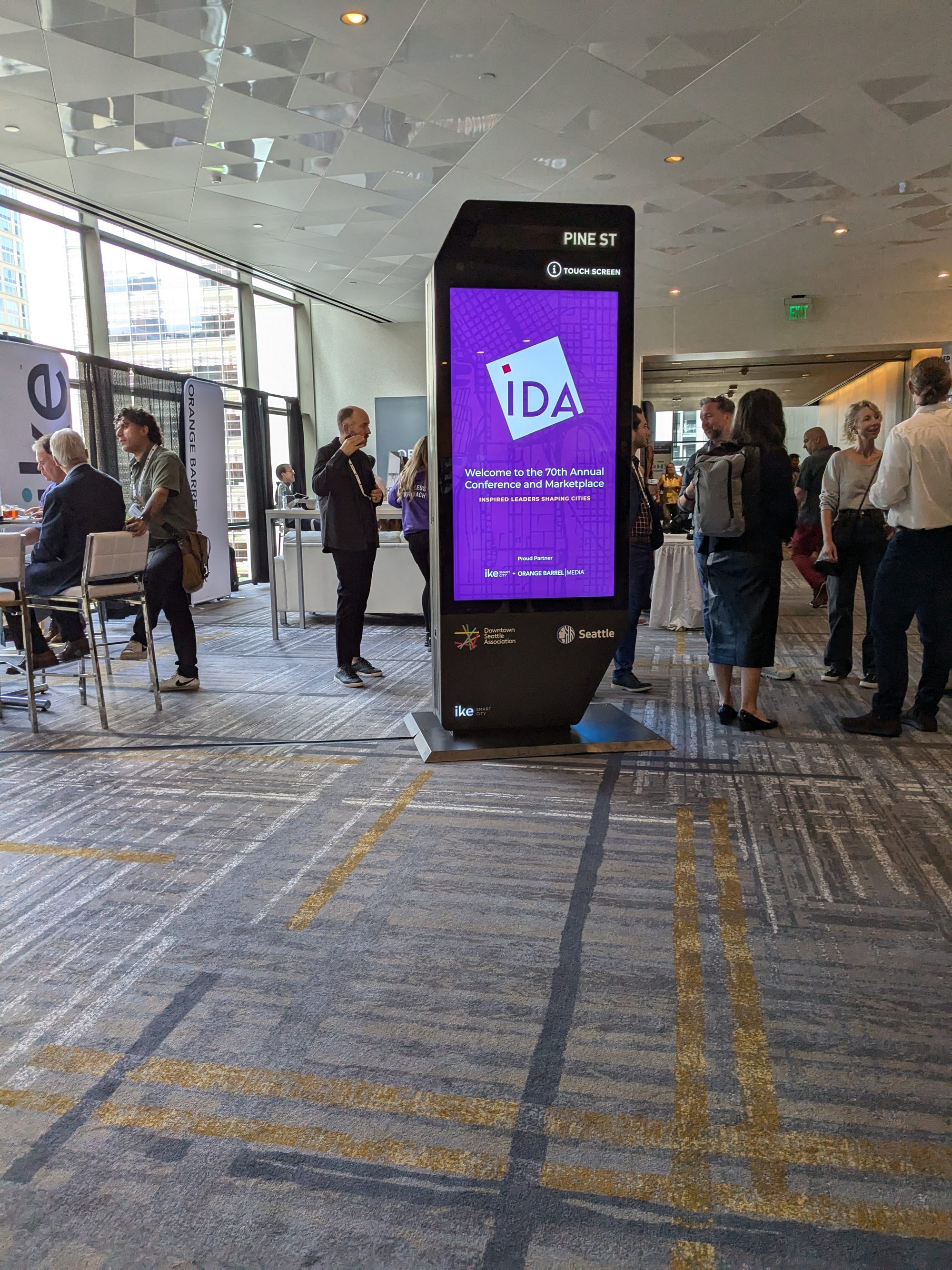 An IKE digital kiosk demo unit labeled PINE ST on the show floor of the International Downtown Association conference in Seattle, September 2024, its screen reading Welcome to the 70th Annual Conference and Marketplace with Proud Partner IKE Smart City + Orange Barrel Media, the Downtown Seattle Association logo and Seattle city seal on its cabinet, and the IKE and Orange Barrel booth banners behind it as attendees mingle.