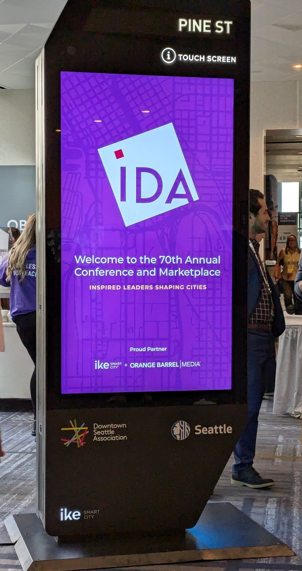 Close-up of the IKE demo kiosk's screen at the conference: the IDA logo above the text Welcome to the 70th Annual Conference and Marketplace, Inspired Leaders Shaping Cities, and below it Proud Partner — IKE Smart City + Orange Barrel Media, with the Downtown Seattle Association logo and Seattle city seal on the cabinet.