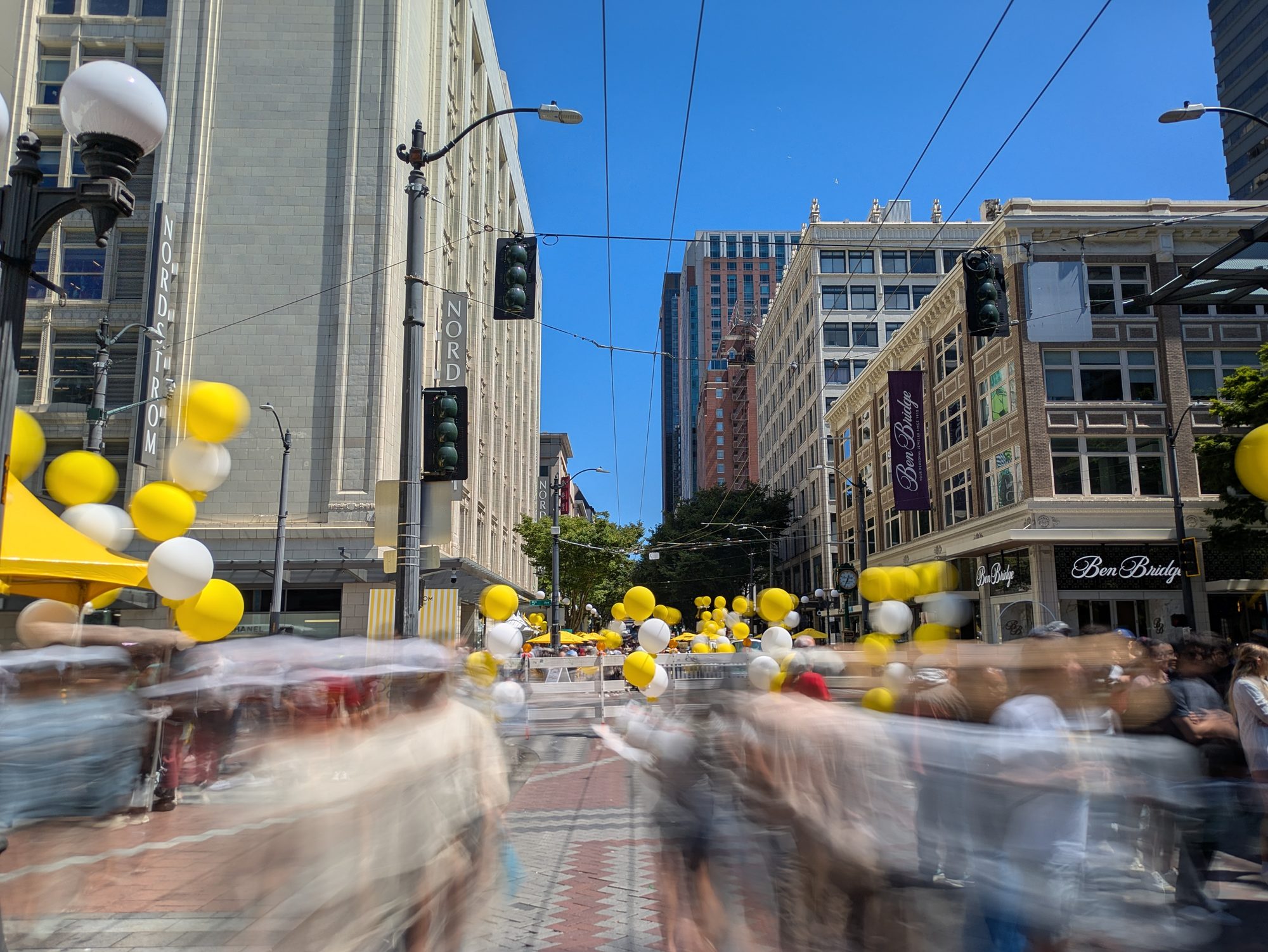 Long-exposure photograph of a closed downtown Seattle street at Pine and 5th during a summer 2026 activation — yellow and white balloons line the street between Nordstrom and Ben Bridge, and the crowd blurs with motion under a clear blue sky.
