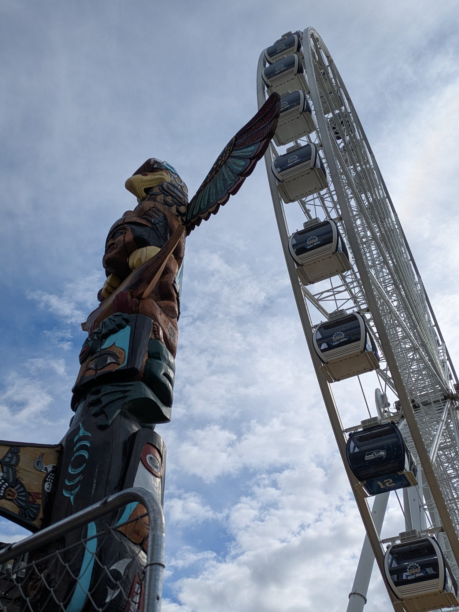 Looking up from the Seattle waterfront at a carved story pole — a winged figure, wings outstretched — beside the white arc of the Seattle Great Wheel, its gondolas climbing into a clouded blue sky. Photo by the author.