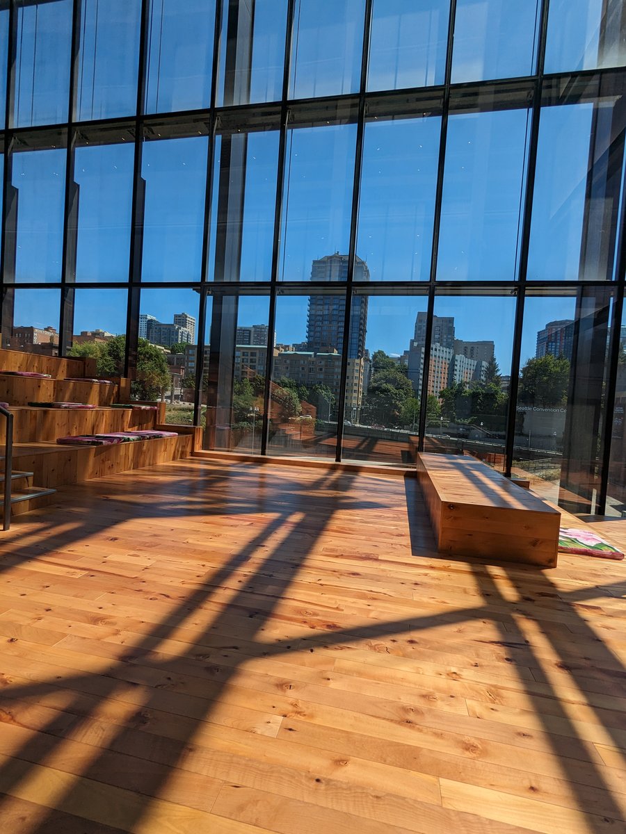 Inside the Summit building's window bay, wooden bleacher steps at left and a low cedar bench at right, the window mullions' shadows raking a warm wood floor, and beyond the glass curtain wall the apartment towers and trees of the neighborhood looking back in under a clear sky. Photo by the author.