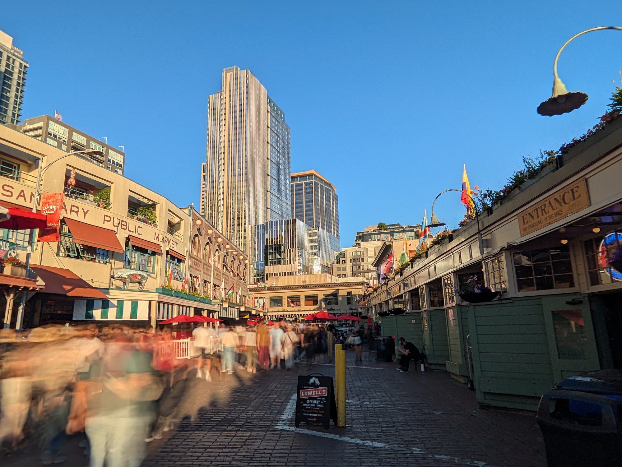 Late afternoon on the cobbles below the Pike Place Market arcade, the Sanitary Public Market building on the left and downtown towers rising behind it. A long exposure turns the passing crowd into a blur while the buildings, a sandwich board and an ENTRANCE sign stay sharp.