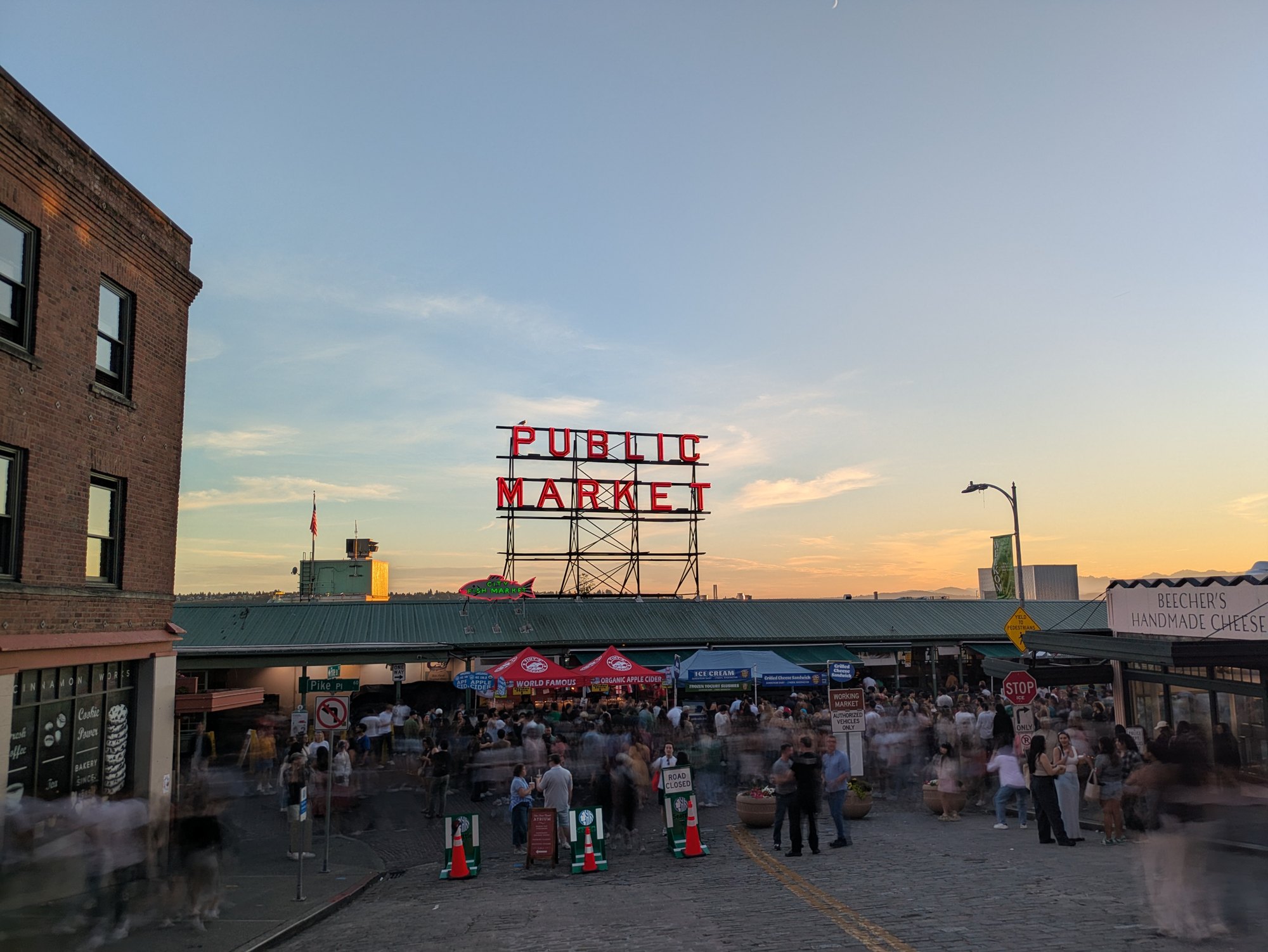 The Pike Place Market sign in red neon at dusk, seen from Pike Street, with the crowd beneath it blurred into streaks by a long exposure while the sign, the stalls and Beecher's Handmade Cheese stay sharp. Photo by the author.