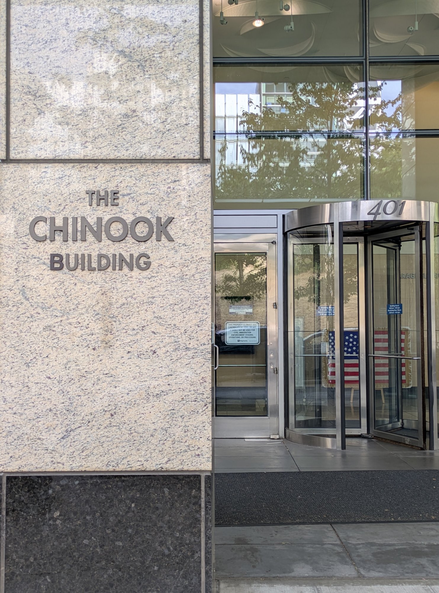 The street entrance of the Chinook Building at 401 Fifth Avenue, King County's office tower in downtown Seattle. The building's name is set in metal letters on a granite pier beside a stainless revolving door, with an American flag standing in the lobby beyond the glass.