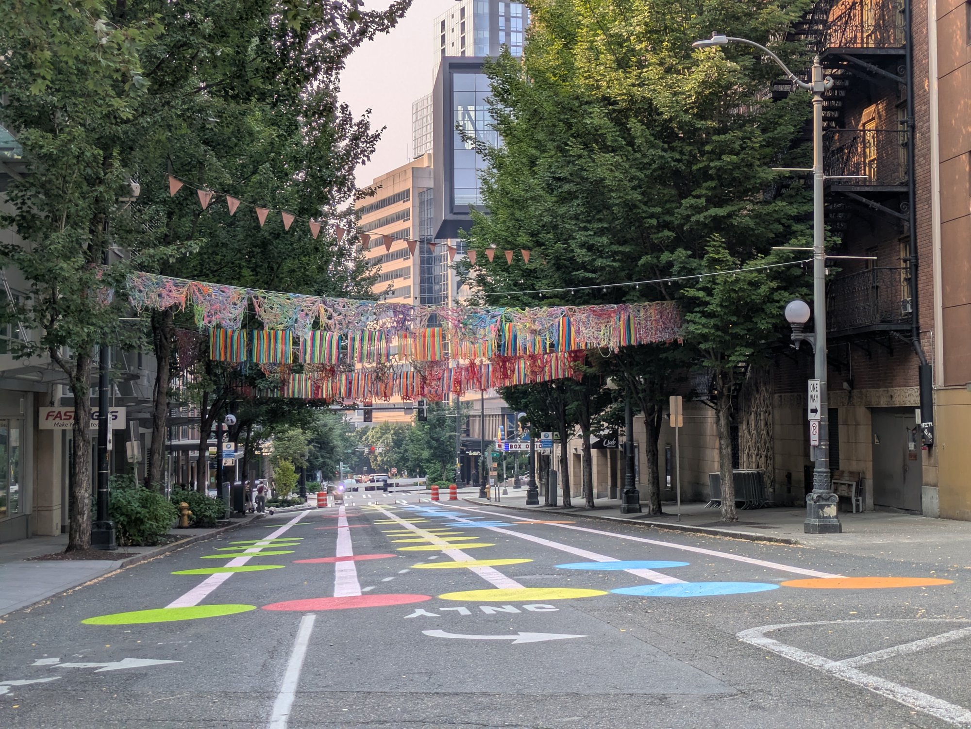 Ninth Avenue closed to cars, looking downhill between Pike and Pine. A canopy of brightly colored knitted and crocheted yarn is strung across the street above the trees, with a line of pennant flags beyond it, and the pavement below is painted with large green, red, yellow, blue and orange ovals over the old lane markings. The block is empty of people and traffic.