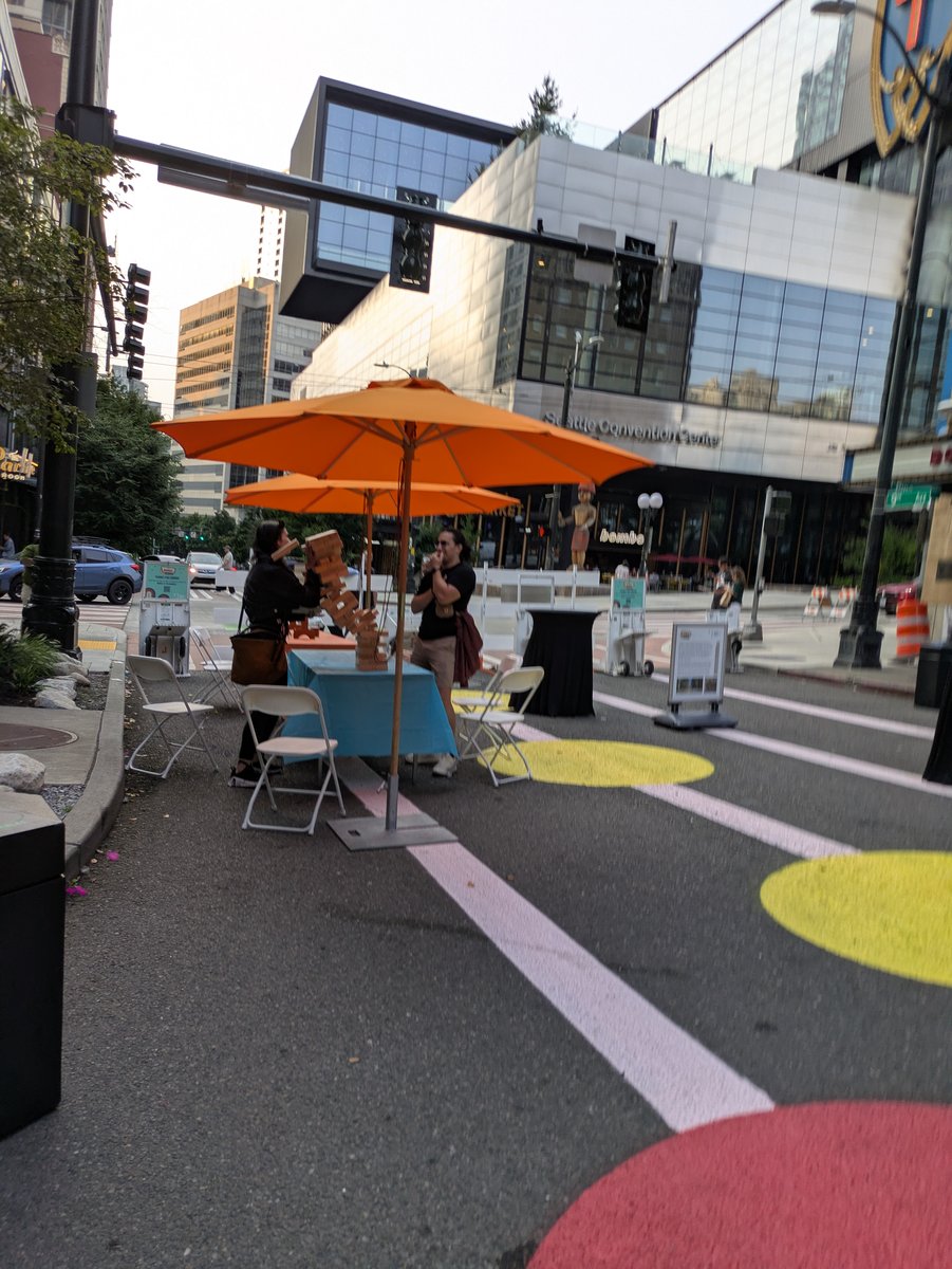 Two people play giant Jenga at a folding table under an orange umbrella on a closed street, standing on pavement painted with large yellow, pink, and red circles and white lines.