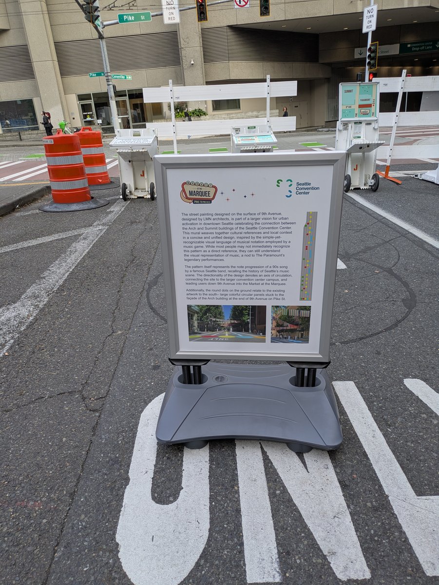 A roadside interpretive sign on 9th Avenue titled 'Market at the Marquee,' with the Seattle Convention Center logo, crediting LMN architects for the street painting design and explaining its meaning, with two photos of the finished pavement art below the text.