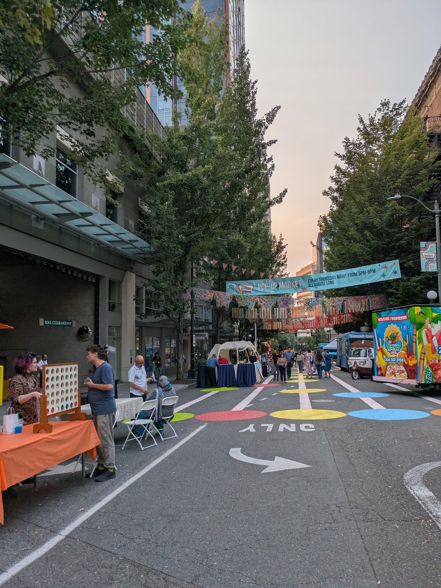 A wide view down 9th Avenue at dusk during Market at the Marquee: a banner reading POP-UP MARKET EVERY THURSDAY NIGHT FROM 5PM-8PM ALL AUGUST LONG hangs over the street, colorful yarn decorations strung below it, food trucks and vendor tables on both sides, and the pavement painted with large red, yellow, blue, and green circles alongside a white lane arrow and the word ONLY.