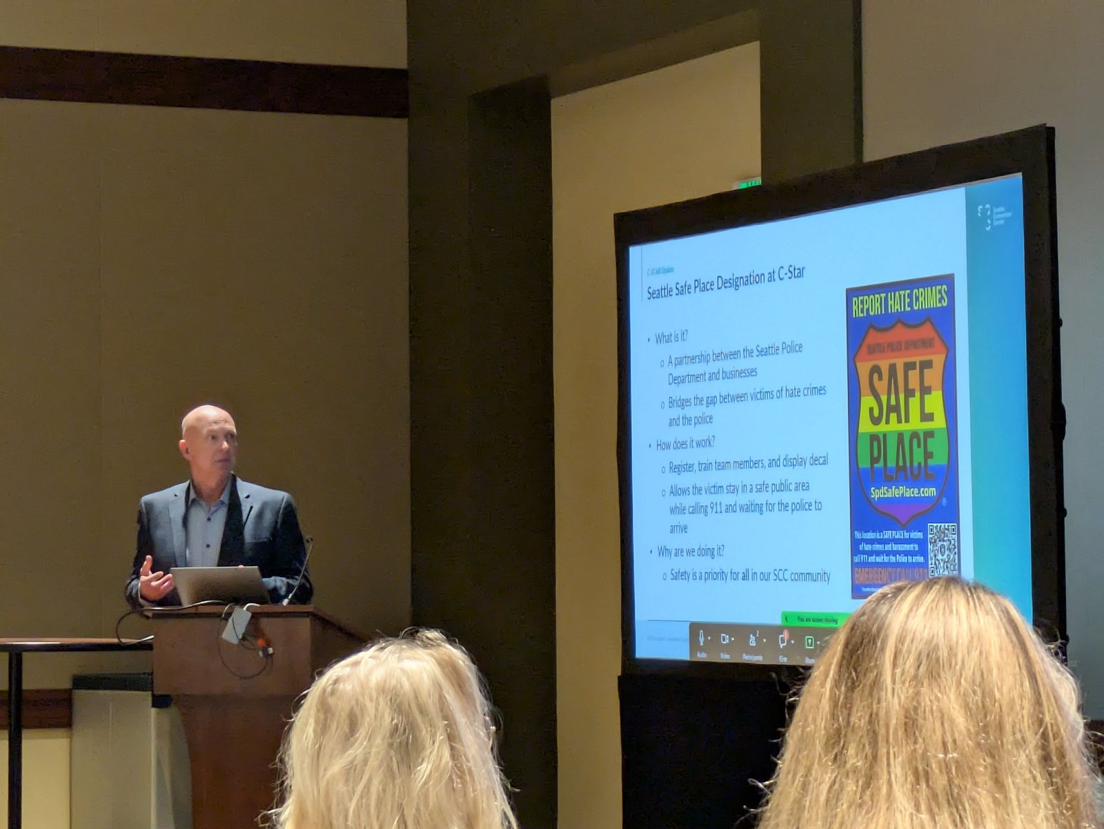 Jesse Anderson, the convention center's executive director of emergency management, speaks at a podium beside a screen reading "Seattle Safe Place Designation at C-Star," with the Seattle Police Department's rainbow Safe Place decal.
