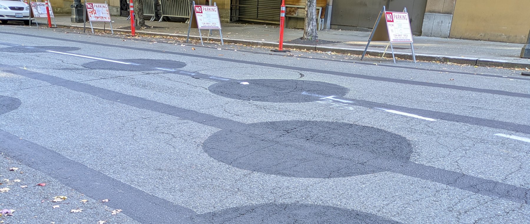 Dark painted-over circles on the pavement of Ninth Avenue, with a curb, red-and-white No Parking signs and orange delineator posts along the sidewalk behind them.